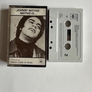 May include: A black and white cassette tape with a silver case. The front of the case features a photo of Johnny Mathis and the text "Johnny Mathis Mathis Is..." and "Including: Sweet Love Of Mine". The back of the case features the text "MATHIS IS Heaven Must Have Made You Just For Me. Hang Up In The Middle Of Love. World Of Laughter. Lullaby Of Love, Sweet Love Of Mine. JOHNNY MATHIS CBS 40-86023".