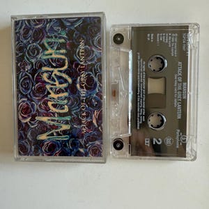 Puede incluir: Una cinta de casete del álbum "Attack of the Grey Lantern" de la banda Mansun. La cinta de casete está en una caja de plástico transparente con una etiqueta negra. La portada del álbum presenta un diseño floral azul oscuro y morado con el nombre de la banda en blanco.