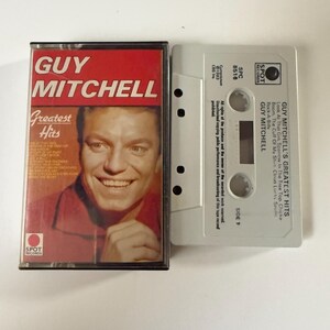 May include: A cassette tape case with a red background and a picture of Guy Mitchell. The text "Guy Mitchell Greatest Hits" is printed on the case. The cassette tape is white with black text.