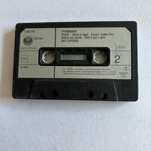 Könnte beinhalten: Eine schwarze Kassette des Def Leppard Albums Pyromania. Das graue Etikett zeigt Songtitel wie "Rock of Ages" und "Billy's Got a Gun". Das Etikett enthält auch das Vertigo-Logo und "Made in Holland". Die Kassette ist Stereo.