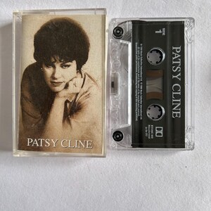 May include: A vintage cassette tape with a black case and a clear plastic cover. The tape features a photo of Patsy Cline on the front with the text "Patsy Cline" printed below the photo. The tape is labelled "Side 1" and "Patsy Cline" on the case.
