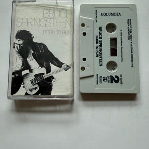 Puede incluir: Un casete de Bruce Springsteen Born to Run con su caja de plástico transparente. La carátula muestra una fotografía en blanco y negro del artista con una guitarra. La carcasa gris claro del casete muestra el logo de Columbia y la lista.