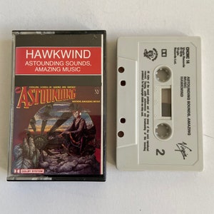 May include: A cassette tape of the album "Astounding Sounds, Amazing Music" by the band Hawkwind. The cassette tape is white with black text and a black Virgin Records logo. The cassette tape is in a plastic case with a picture of a woman sitting on a large metal wheel.