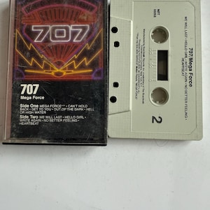 Peut inclure: Une cassette audio vintage de l'album "707 Mega Force". Le boîtier de la cassette est noir avec un design néon et le chiffre 707. La cassette elle-même est blanc cassé avec le titre de l'album et la liste des titres.
