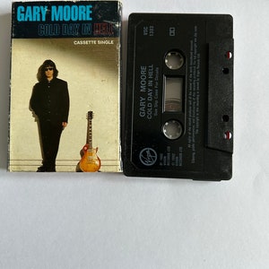 Pode incluir: Uma fita cassete preta com o título "Gary Moore - Cold Day in Hell" e o texto "See Slip Case For Details". A fita cassete está sobre um fundo branco.