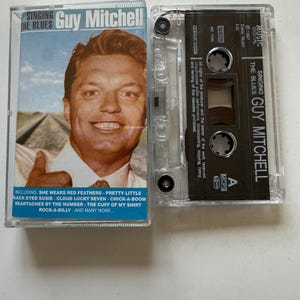 May include: A vintage cassette tape of Guy Mitchell's album "Singing the Blues." The cassette case is clear, with the album cover featuring a smiling man in a white shirt. The tracklist includes "She Wears Red Feathers" and "Rock-A-Billy."