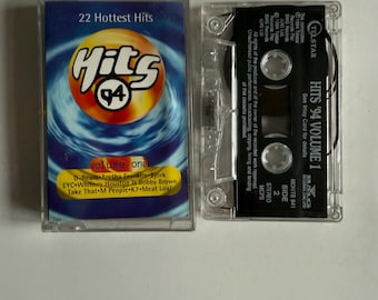 Może przedstawiać: Przezroczysty plastikowy futerał na kasetę magnetofonową z czarną kasetą w środku. Kaseta jest oznaczona jako "Hits '94 Volume 1" i ma czarno-biały projekt. Futerał ma niebiesko-białe tło z tekstem "22 Hottest Hits" i "Hits '94" w białym kółku z czarną cyfrą 4. Tekst "Volume One" znajduje się pod kółkiem i wymienia artystów, którzy są na kasecie.