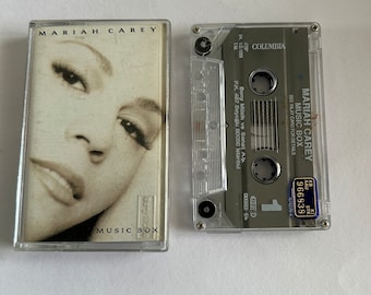 May include: A clear plastic cassette tape case with a black cassette tape inside. The cassette tape is labeled "Mariah Carey Music Box" and has a picture of Mariah Carey on the front. The cassette tape is a Columbia Records release.