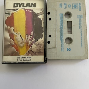 May include: A vintage cassette tape with a black and white photo of Bob Dylan on the front. The tape is labeled "DYLAN" and features the songs "Lilly Of The West" and "A Fool Such As I".