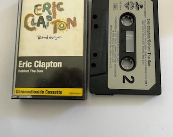 Puede incluir: Una cinta de casete del álbum "Behind the Sun" de Eric Clapton. La cinta de casete es negra con una etiqueta plateada que dice "Eric Clapton/Behind The Sun" y "2". La cinta de casete está en una caja de plástico transparente con una etiqueta blanca que dice "Eric Clapton" y "Behind the Sun". La etiqueta también dice "Chromedioxide Cassette".
