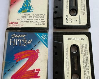 Może przedstawiać: Cztery zabytkowe kasety z serii Super Hits. Obudowy kaset mają kolorowe wzory z numerami 1 i 2 w dużych czcionkach. Kasety są czarne z białymi etykietami, a tekst "Superhits #1" i "Superhits #2" jest widoczny.