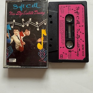 May include: A vintage cassette tape of the Soft Cell album "Non-Stop Erotic Cabaret." The cassette case features a black background with the band's name and album title in blue and pink. The cassette itself is black with pink labels.