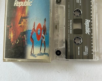 Könnte beinhalten: Ein transparentes Plastik-Kassettengehäuse mit einem schwarzen Kassettenband im Inneren. Das Kassettenband trägt den Text "A New Order Release" und "Republic". Das Kassettenband ist eine Musikalbenveröffentlichung.