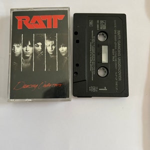 Op de afbeelding: Een zwarte cassetteband met de titel "RATT/DANCING UNDERCOVER" en de songtitels "DANCE", "ONE GOOD LOVER", "DRIVE ME CRAZY", "SLIP OF THE LIP", "BODY TALK". De cassetteband bevindt zich naast een zwart-witfoto van een band met de titel "RATT" en "Dancing Undercover" in rood.