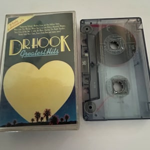 May include: A cassette tape case with a blue background and a heart-shaped image of a sunset with palm trees. The case has the text "Dr. Hook Greatest Hits" on the front. The cassette tape is visible in the case.
