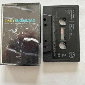 Może przedstawiać: Czarna kaseta magnetofonowa z białą etykietą. Etykieta zawiera tekst "OMD SUGAR TAX" i "ORCHESTRAL MANOEUVRES IN THE DARK".