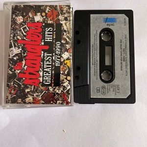 Puede incluir: Una cinta de cassette negra con el título "The Stranglers Greatest Hits 1977-1990" y la etiqueta "epic".