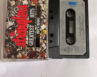 Może przedstawiać: Czarna kaseta magnetofonowa z tytułem "The Stranglers Greatest Hits 1977-1990" i etykietą "epic".