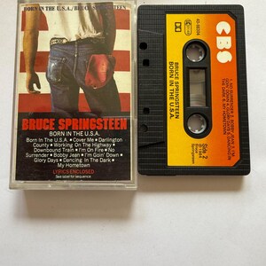 Puede incluir: Un casete de Bruce Springsteen Born in the U.S.A. con su caja de plástico. La carátula muestra la imagen de una persona con vaqueros y camisa blanca sobre un fondo de bandera. El casete tiene una etiqueta amarilla y naranja con la lista de temas.