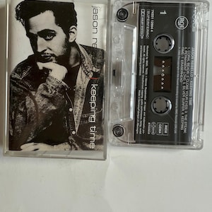 Può includere: Una musicassetta trasparente con un'immagine in bianco e nero di un uomo. L'etichetta riporta "jason re keeping time" e include la tracklist dell'album e le informazioni sull'artista.