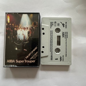May include: A white cassette tape with a black label. The label has the text "ABBA Super Trouper" and "The Gift of Music" with a smaller text "Win a Saab". The cassette tape is in a plastic case with a picture of the band ABBA on the front.