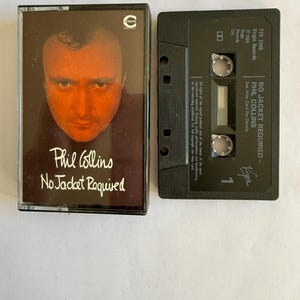 Peut inclure: Une cassette audio vintage de l'album "No Jacket Required" de Phil Collins. L'étui de la cassette présente un portrait de Phil Collins et le titre de l'album en texte blanc. La cassette noire est à côté de l'étui.