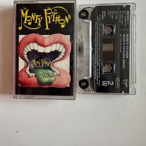 Può includere: Una cassetta con un'immagine in bianco e nero di una bocca con la lingua che sporge. Il testo "Monty Python Sings" è sulla cassetta.