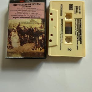 Op de afbeelding: Twee vintage cassettes. Eén is zwart met een kleurrijk label met tekst en een illustratie van mensen. De andere is lichtgeel met tekst en het nummer 2. Beide cassettes zijn voor klassieke muziek.