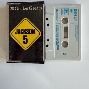 May include: A cassette tape case with a black background and a yellow diamond with the text "JACKSON 5" inside. The case has the text "20 Golden Greats" on the front. The cassette tape is white with blue text and a blue and white label.