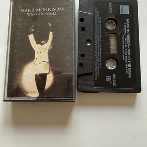 Puede incluir: Una cinta de cassette y su estuche de Mark Morrison, "Who's The Mack!". El estuche presenta una imagen en blanco y negro de una persona con los brazos levantados. La cinta es negra con texto blanco.