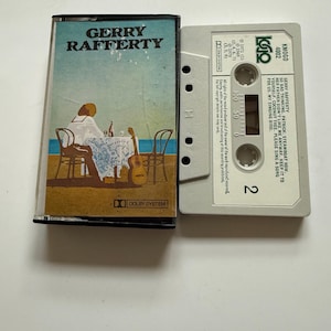 Peut inclure: Une cassette audio vintage avec l'album "Gerry Rafferty". L'étui de la cassette présente une illustration colorée d'une personne à une table avec une guitare. La cassette est grise claire avec du texte imprimé.