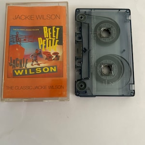 Peut inclure: Une cassette audio avec un boîtier en plastique transparent et une étiquette noire. La cassette audio est à côté d'un boîtier de cassette audio avec un fond orange et une image d'un personnage de dessin animé. Le texte sur le boîtier indique "Jackie Wilson, The Classic Jackie Wilson, Reet Petite".