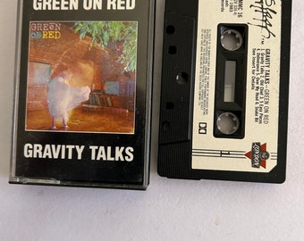 May include: A black cassette tape with a white label. The label has the text "Green on Red" and "Gravity Talks" in large white letters. The cassette tape is in a plastic case with a black background and a picture of a person in a white shirt standing in front of a brick wall.