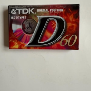 Puede incluir: Un paquete de cinta de cassette rojo, negro y dorado con el texto "TDK. NORMAL POSITION 120/μSEC IEC I/TYPE I D 60" en la parte delantera.