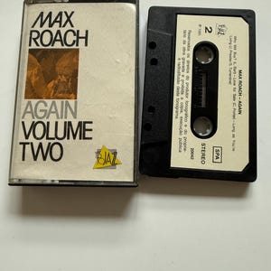 May include: A vintage cassette tape of Max Roach's album "Again Volume Two." The cassette case is clear with black text and a yellow logo. The cassette itself is black with white text, including the album title and song information. The album was released in 1985.