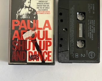 May include: A black cassette tape with a white label. The label has the text "PAULA ABDUL SHUT UP AND DANCE (THE DANCE MIXES)" and a Virgin Records logo. The cassette tape is on a white surface.