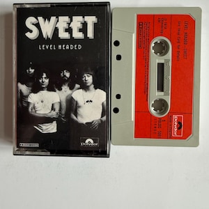 Puede incluir: Una cinta de cassette vintage del álbum "Level Headed" de la banda Sweet. La caja negra de la cinta muestra el nombre de la banda y el título del álbum en blanco. La cinta es gris con una etiqueta roja. La portada presenta una foto en blanco y negro de la banda.