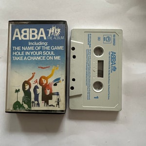 Op de afbeelding: Een witte cassetteband met een zwart label. Het label heeft de tekst "ABBA" in blauw en wit, en de tekst "The Album" in kleinere witte letters. De cassetteband is gelabeld met de titel "The Name of the Game" en bevat de nummers "Hole in Your Soul" en "Take a Chance on Me".