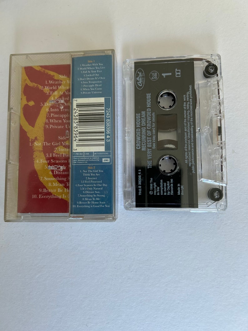 May include: A cassette tape with a clear plastic case and a black label. The label has the text "Crowded House Recurring Dream The Very Best of Crowded House" printed on it. The cassette tape is a music album.