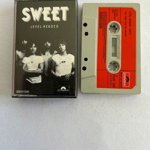 Puede incluir: Una cinta de casete del álbum "Level Headed" de la banda Sweet. La cinta de casete es roja y blanca con el nombre de la banda y el título del álbum impreso en ella. La cinta de casete está en una caja de plástico con una foto en blanco y negro de la banda en la parte delantera.