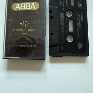 Op de afbeelding: Een ABBA Dancing Queen cassette single met bijbehorend zwart plastic doosje. De hoes toont de bandnaam in goud, de titel en het nummer Lay All Your Love On Me. De zwarte cassette toont de tracklist en de logo's van Polydor en Polar.