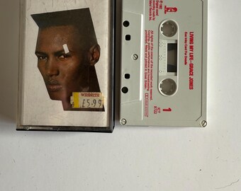 May include: A cassette tape of Grace Jones's album "Living My Life". The cassette tape is white with red text and a red barcode. The album cover is a close-up of Grace Jones's face. The album cover is black and white with a red price tag.