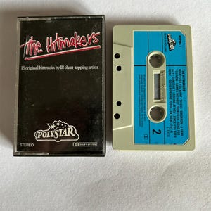 Könnte beinhalten: Eine Kassette mit einem blauen Etikett und dem Text "The Hitmakers" und "18 original hit tracks by 18 chart-topping artists". Die Kassette ist mit "Polystar" beschriftet und hat ein Dolby-System-Logo.