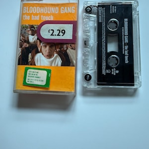 Peut inclure: Une cassette audio et son boîtier pour l'album "The Bad Touch" de Bloodhound Gang. Le boîtier orange présente une photo du groupe et du texte. La cassette est transparente avec des détails et du texte noirs.