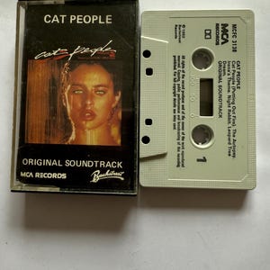 May include: A vintage cassette tape of the "Cat People" original soundtrack. The cassette case features a woman's portrait with the title in bold letters. The cassette tape is light gray with the track list and "MCA Records" printed on it.