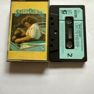 Peut inclure: Une cassette audio vintage et son étui pour la bande originale du film "Sweet Dreams". L'étui est jaune avec le titre du film en bleu et une photo de deux personnes. La cassette est noire avec une étiquette bleu clair.