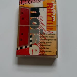 Peut inclure: Une cassette audio vintage avec le texte "Underground + Rhythm" en rouge et jaune. L'étui en plastique transparent affiche une liste de groupes tels que Depeche Mode et Sonic Youth.