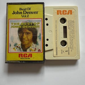 May include: A retro cassette tape of "Best of John Denver Vol. 2" with a yellow and red cover featuring a photo of John Denver. The cassette is open, showing the tape and the RCA logo.