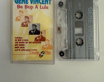 May include: A cassette tape with a clear plastic case and a grey label. The label has the text "GENE VINCENT" and "Be Bop A Lula" printed on it. The cassette tape includes the songs "Be Bop A Lula", "The Story of the Rockers", "Bay Mama", "Baby Blue", and 15 other tracks.