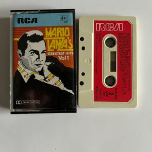 Pode incluir: Uma caixa de cassete preta com um rótulo vermelho. O rótulo tem o texto "RCA" em branco, "Mario Lanza's Greatest Hits Vol 1" em branco, e uma imagem de Mario Lanza.